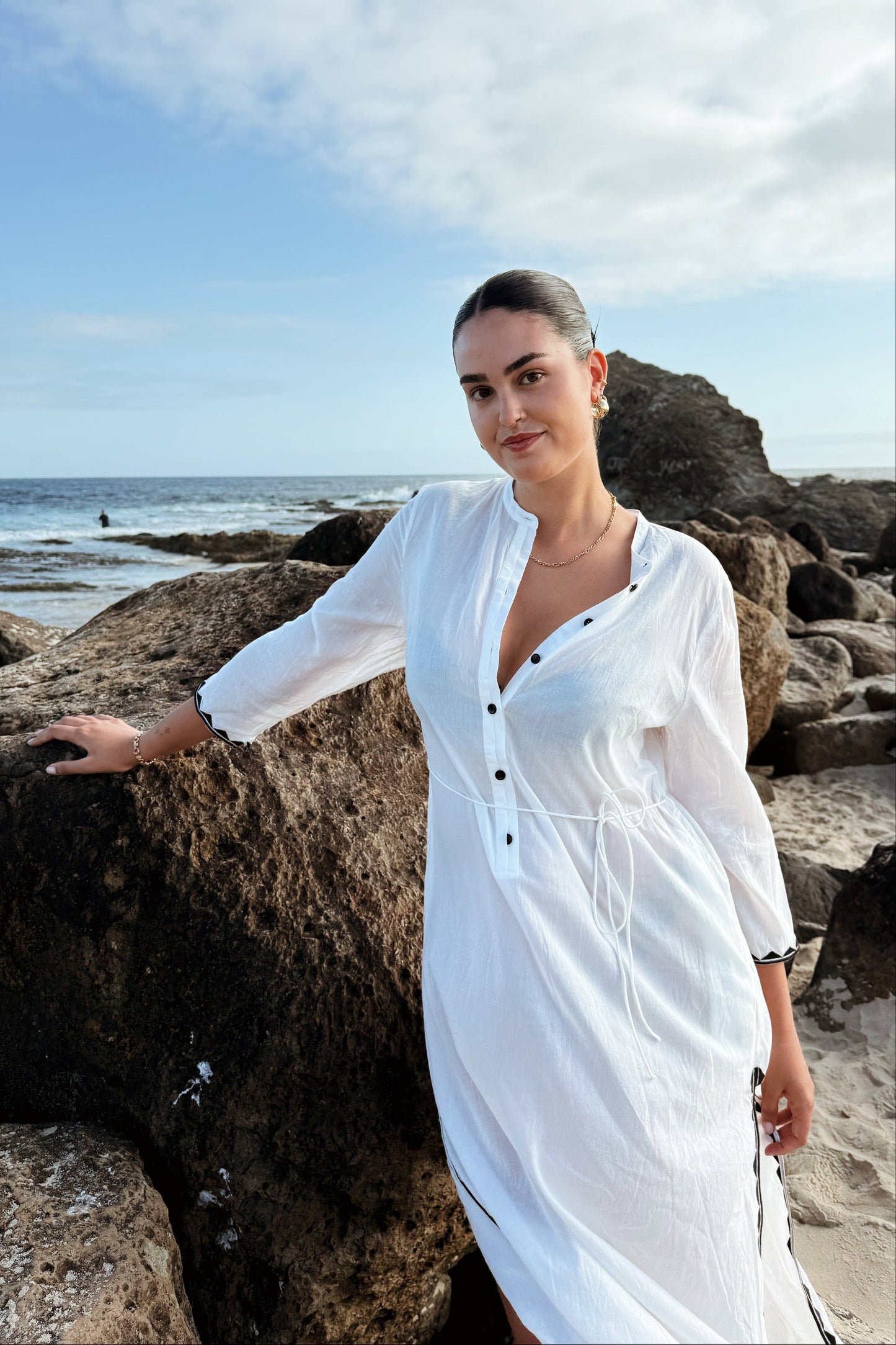 Woman in a white dress standing on a rocky beach with ocean and sky in the background